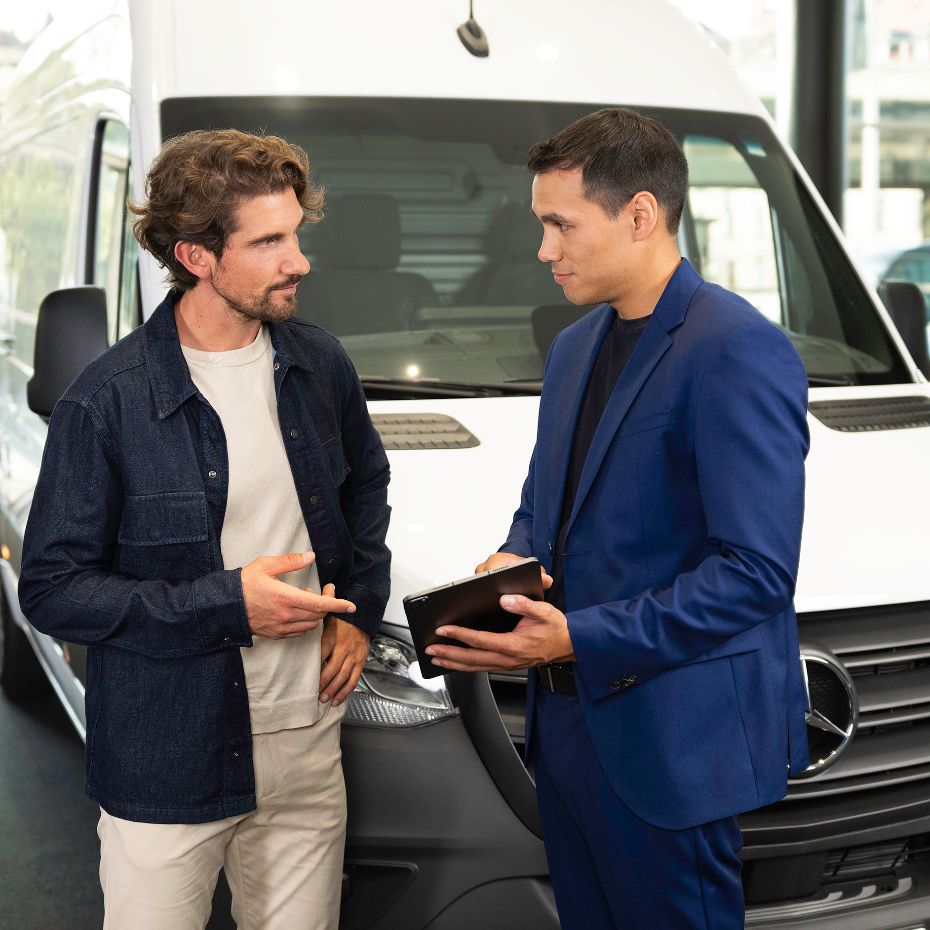 Suscripción Mercedes-Benz para tu Mercedes-Benz Dos personas hablan y señalan con un dispositivo móvil mientras están delante de una furgoneta Mercedes-Benz.